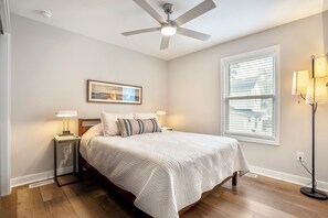 Quarto com cama, luminárias de cabeceira, ventoinha no teto, janela com persianas e quadro na parede.