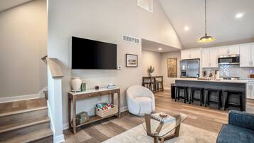A modern kitchen with a large island, a flat-screen TV mounted on the wall, and a dining area with a sofa and a coffee table.