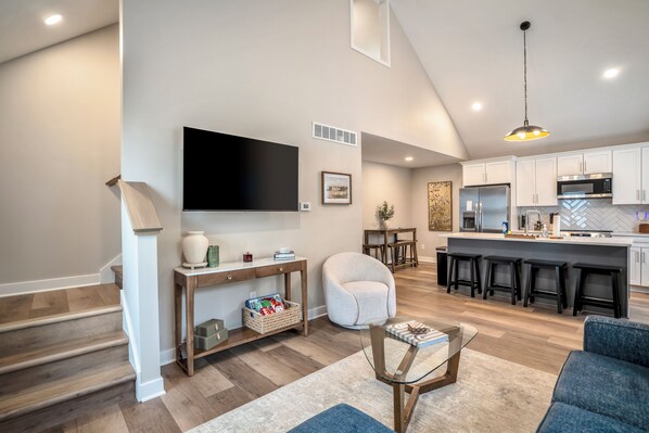 A modern kitchen with a large island, a flat-screen TV mounted on the wall, and a dining area with a sofa and a coffee table.
