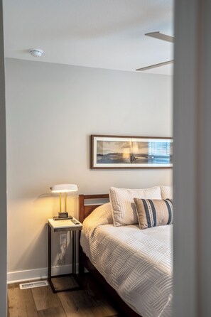 A bedroom with a bed, a bedside table with a lamp, a framed picture on the wall, and a window. - Stay close to your student in this private, brand-new home near campus and downtown Bowling Green, Ohio. Walkable to great dining, enjoy modern comfort, and relax in a peaceful, thoughtfully designed space. (Bowling Green)
