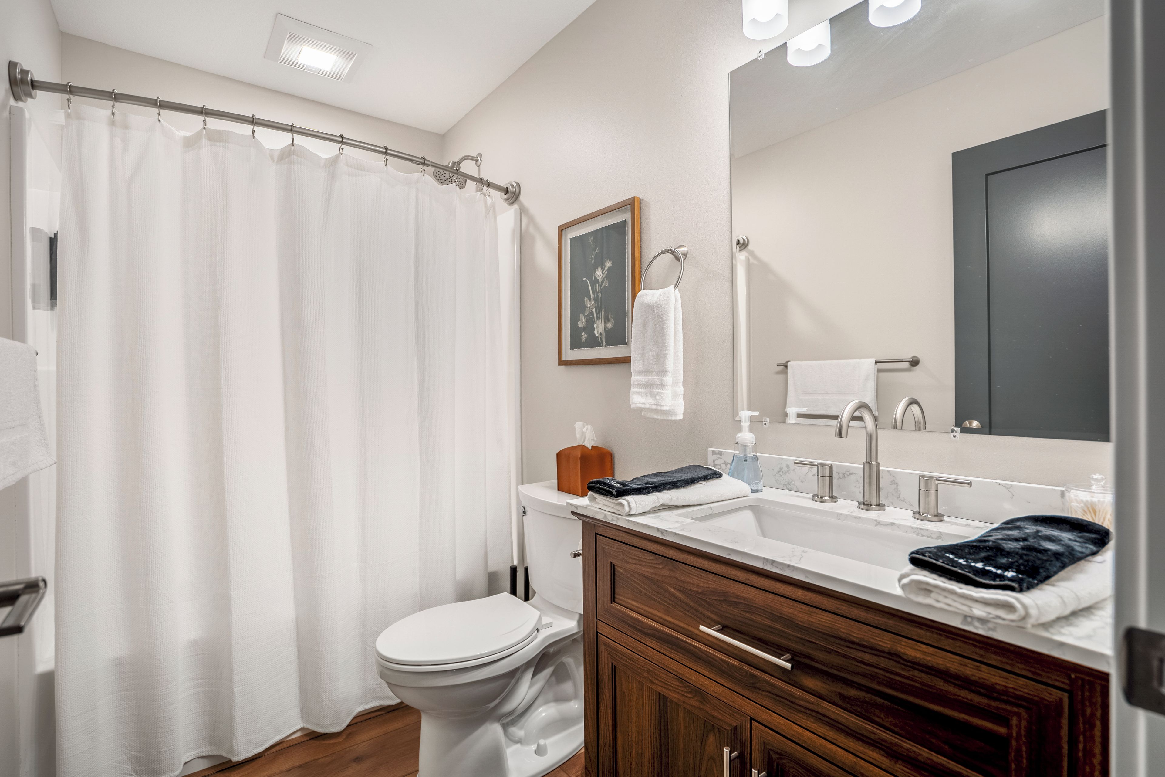 A bathroom with a shower curtain, toilet, sink with a marble countertop, wooden vanity, and a large mirror.