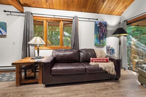 A cozy living room with a brown leather sofa, a wooden table with a lamp, and a window with curtains. - Petros Place Ptarmigan Village Hot Tub Pool (Whitefish)