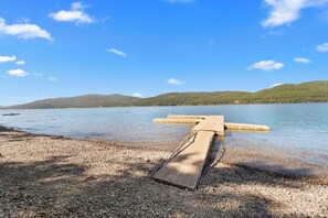 A pebble beach leading to a pier extending into calm waters, surrounded by forested hills. - Petros Place Ptarmigan Village Hot Tub Pool (Whitefish)