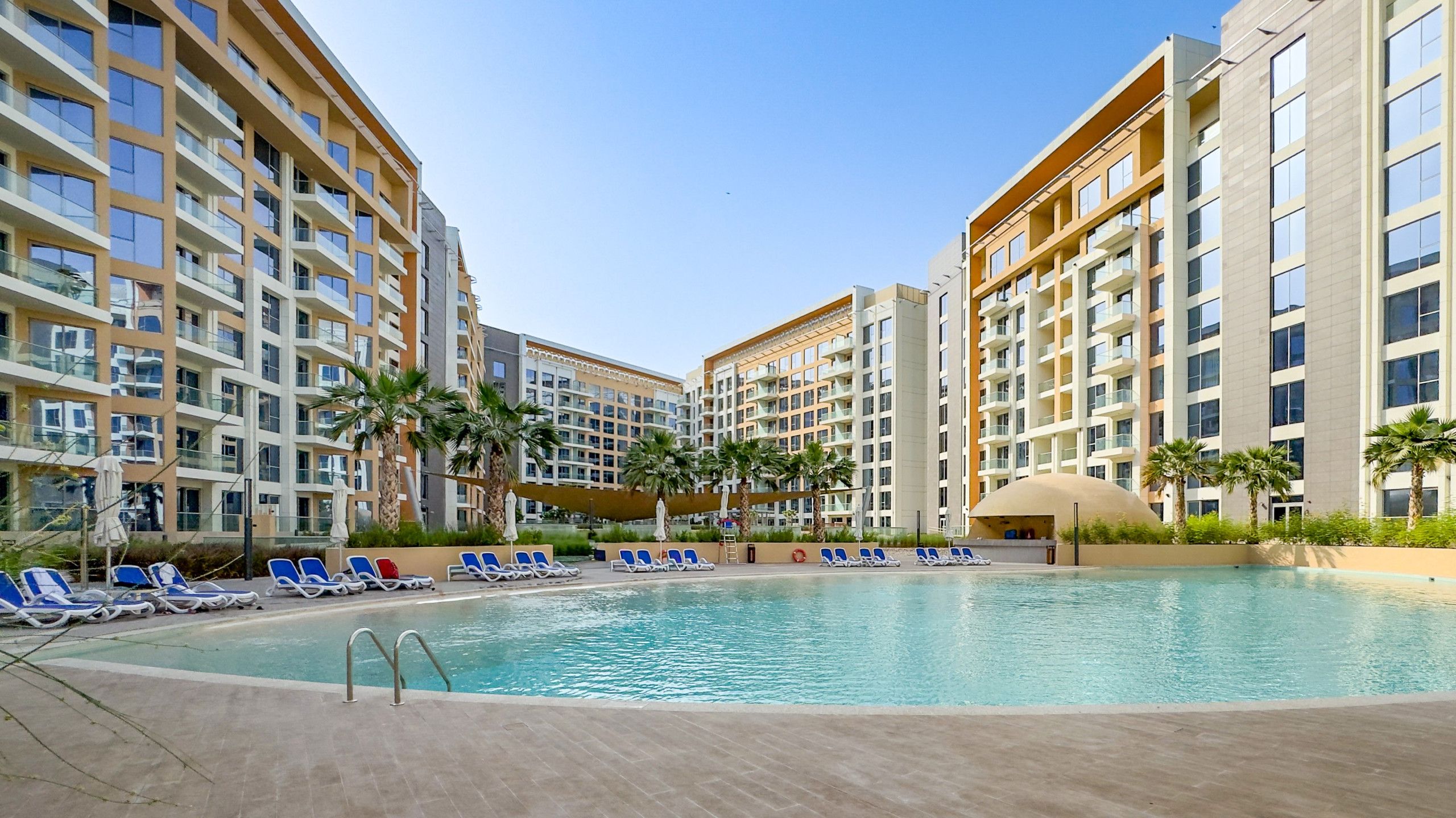 A swimming pool surrounded by modern apartment buildings with balconies, palm trees, and sun loungers.