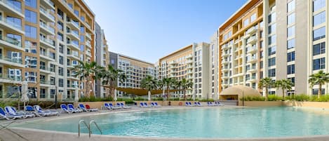 A swimming pool surrounded by modern apartment buildings with balconies, palm trees, and sun loungers.