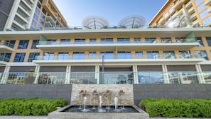 Un hotel de varios pisos con un diseño moderno, que presenta una fuente central con múltiples fuentes, balcones con barandillas de vidrio y un cielo despejado.