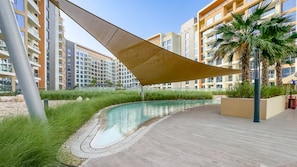 Zona de piscina exterior moderna amb disseny corbat, envoltada per un solàrium de formigó i una vela ombreig gran.