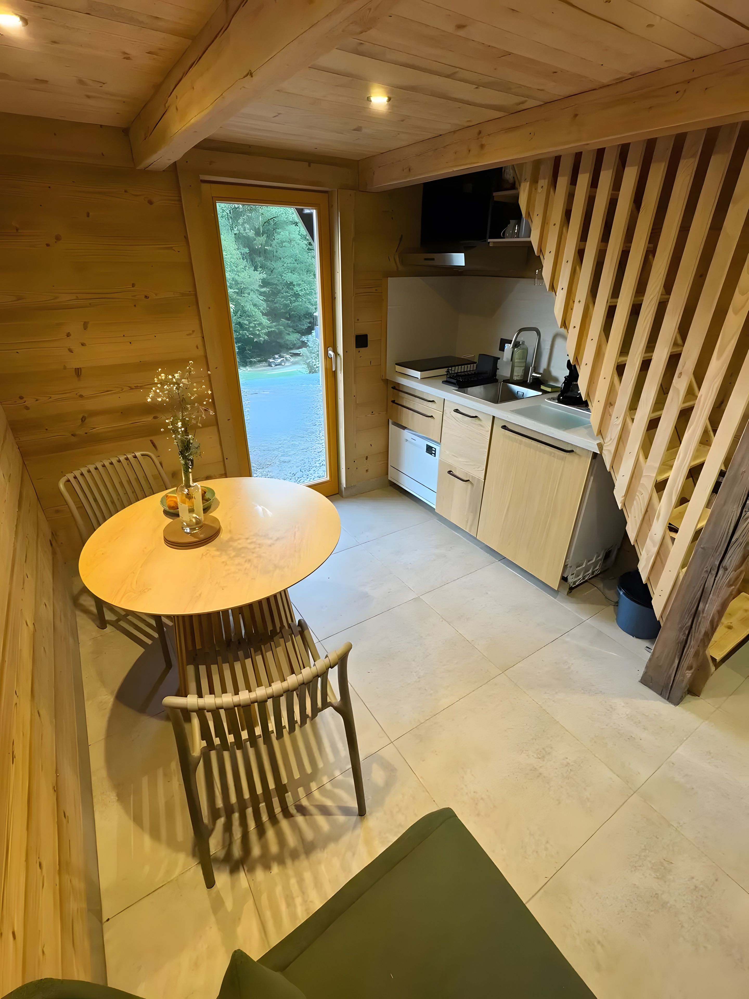 Een houten interieur met een ronde tafel en stoelen, een keuken met kasten en apparatuur, en een uitzicht door een glazen deur.
