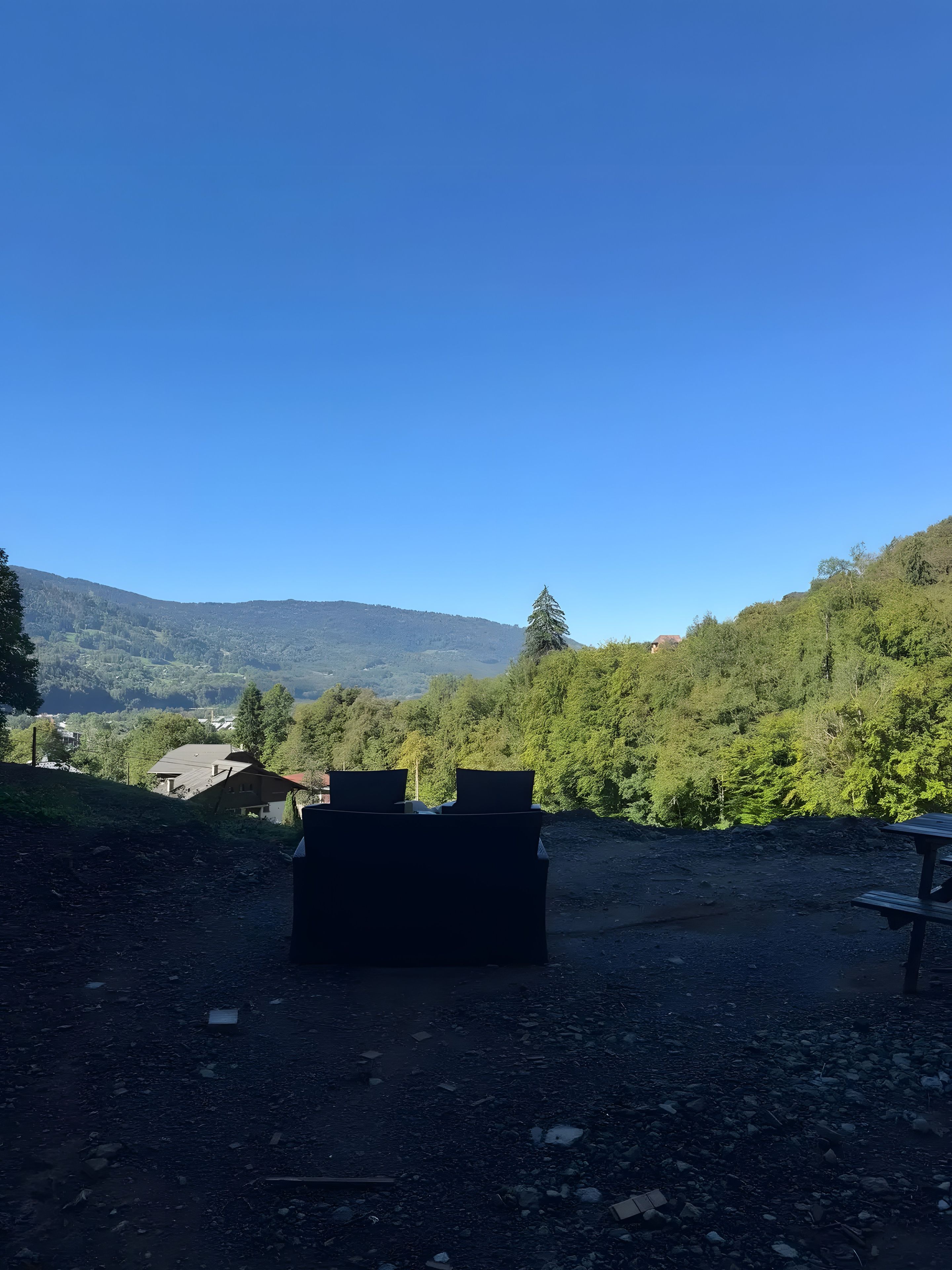 Un espacio con grava, con un banco y una mesa de picnic, rodeado de follaje y colinas al fondo.
