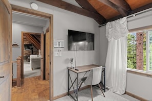 Una habitación con escritorio, silla y un amplio ventanal con cortinas. Hay un televisor montado en la pared y una puerta que da a otra habitación.