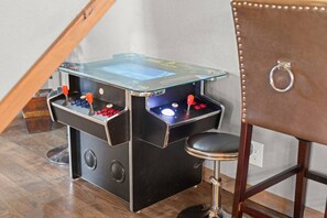 Ein kleiner, abgeschlossener Spielautomaten-Tisch mit zwei Joysticks und einem Münzschlitz, platziert in einer Ecke mit einem Holzstuhl in der Nähe.