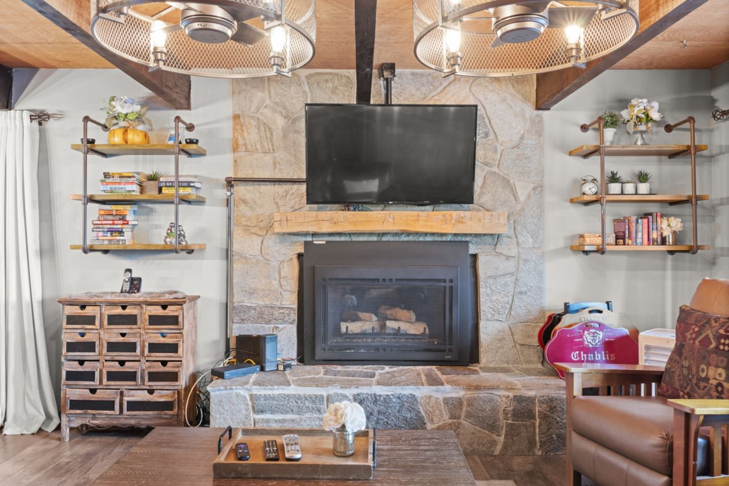 A living room with a stone fireplace, a flat-screen TV mounted above it, wooden shelves with books and decorative items, and a leather couch.