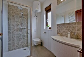 Casa de banho com cabine de chuveiro de vidro, sanita, lavatório com espelho e janela com planta em vaso.