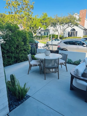 Een terras buiten met een ronde tafel en stoelen, omringd door groen en een straatbeeld.