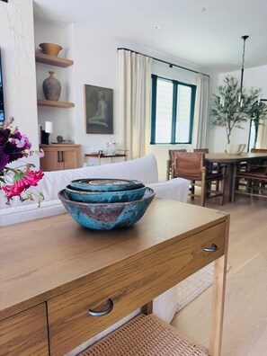 Ein Holztisch mit einer blauen Keramikschale, ein weißes Sofa, ein Essbereich mit Stühlen, ein Gemälde an der Wand und ein Fenster mit Vorhängen.