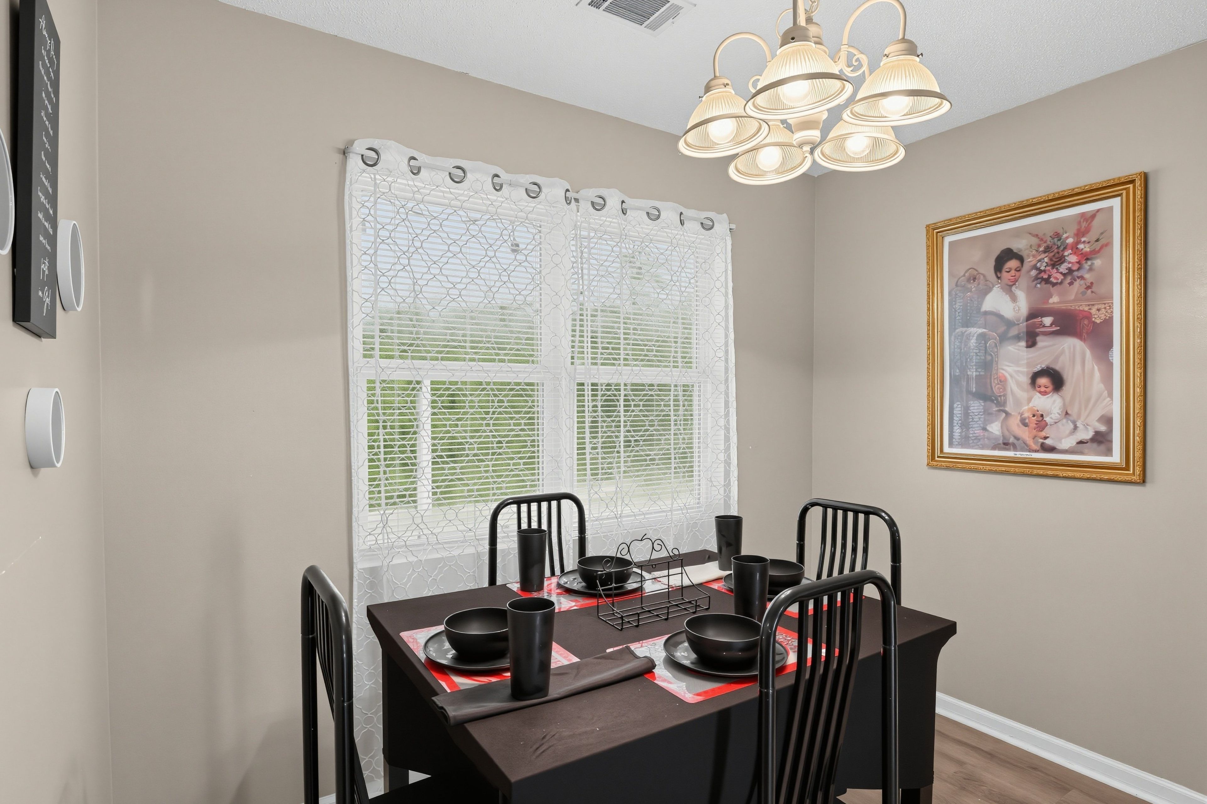A dining area with a table set for four, chairs, a decorative window with a white curtain, a wall-mounted light fixture, and a framed picture.