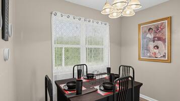 A dining area with a table set for four, chairs, a decorative window with a white curtain, a wall-mounted light fixture, and a framed picture.