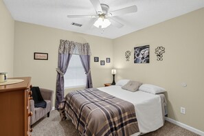 A bedroom with a bed, a nightstand, a lamp, a ceiling fan, and a window with curtains. - Hephzibah Unique Comfortable 4br Retreat For 8 4 Bedroom Home (Hephzibah)