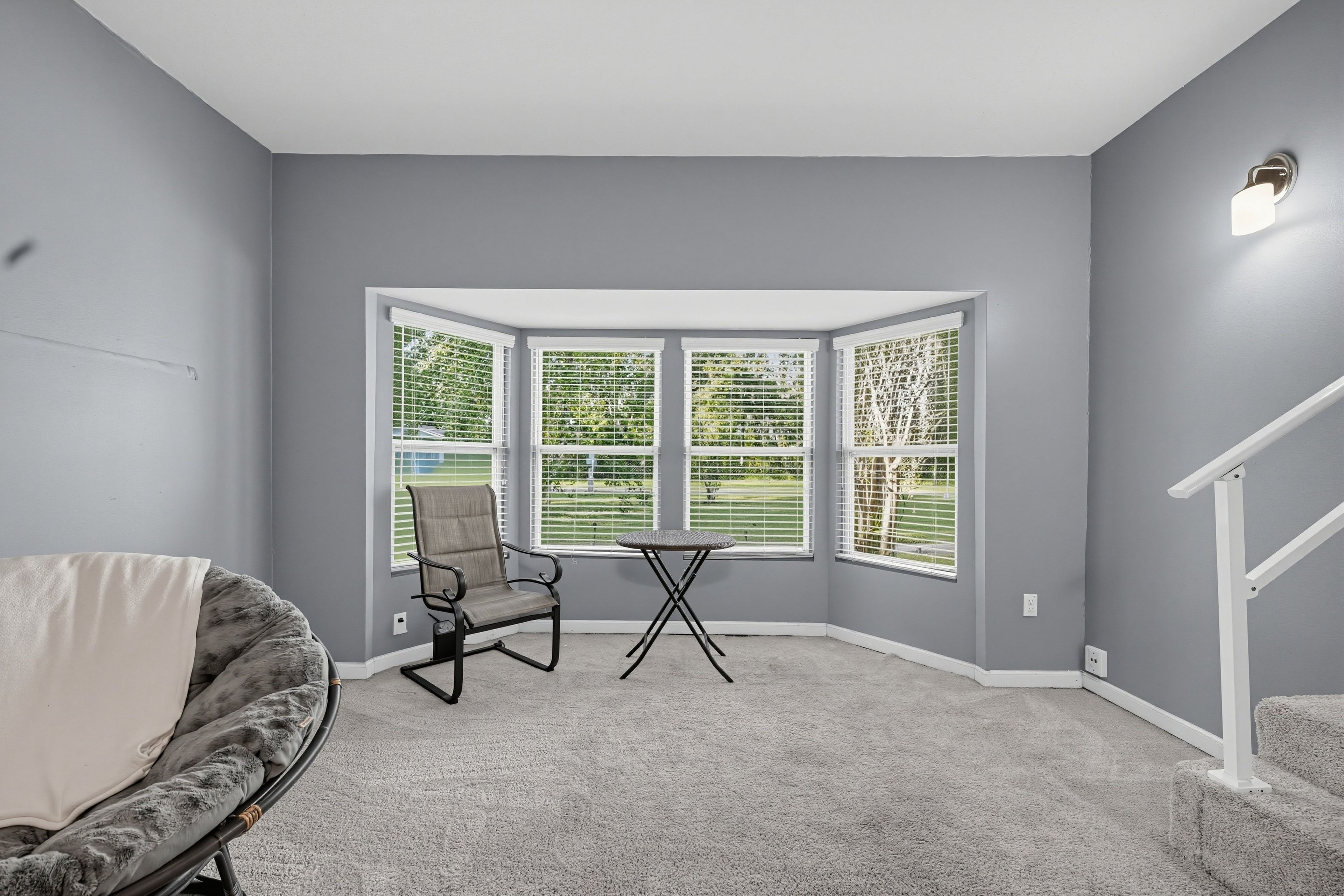 A room with grey walls, a corner window, a grey chair, a small table, and a white staircase railing.