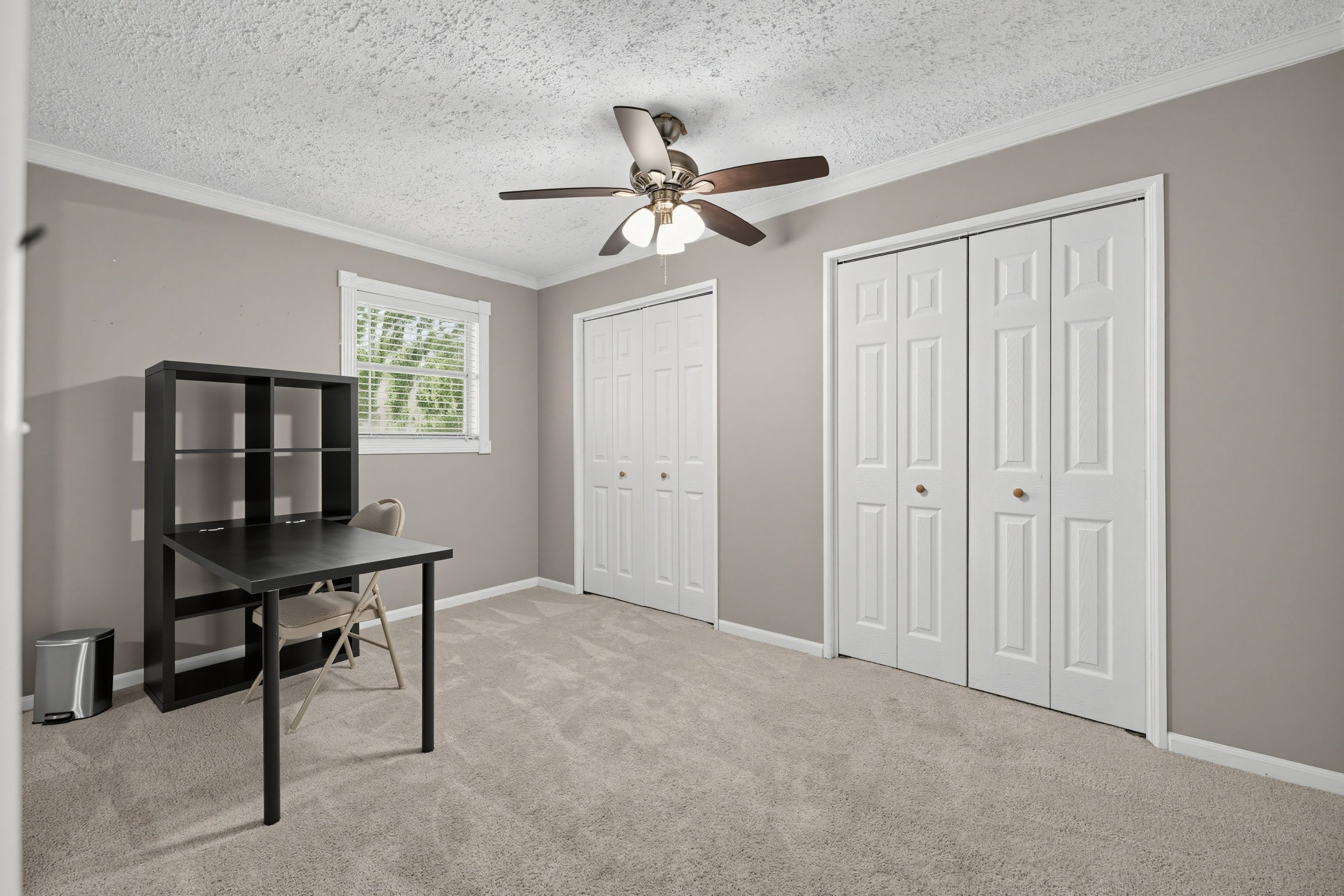 A room with a ceiling fan, a window with blinds, a desk with a chair, and two white doors.