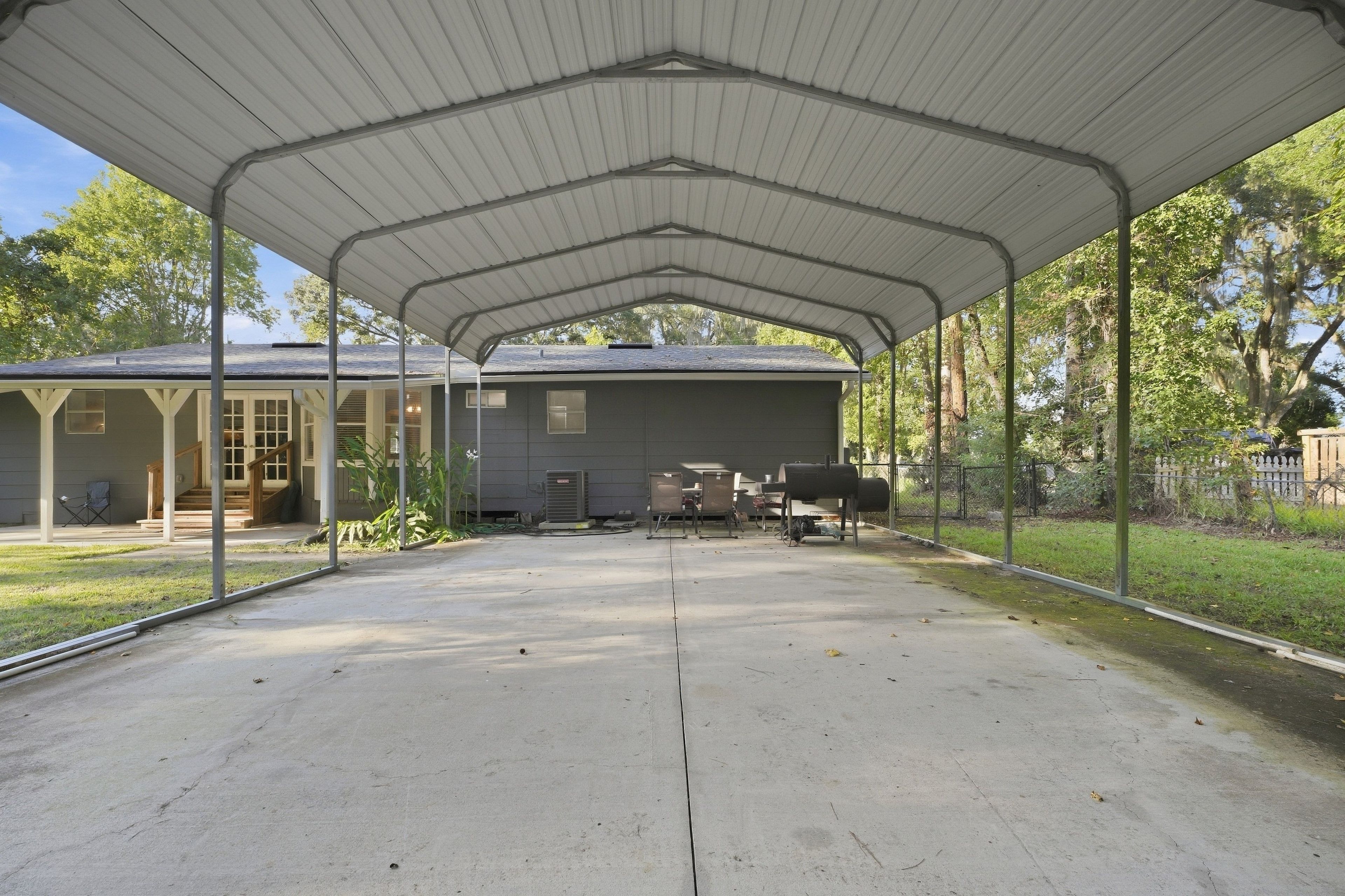 A covered outdoor area with a concrete floor, a grill, and a house in the background.