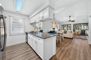 A modern kitchen with white cabinetry and a marble countertop, a dining area with a sofa and chairs, and a large window providing a view of the outdoors. - 146-3 Par For Pawleys 2 Bedroom Condo (Pawleys Island)