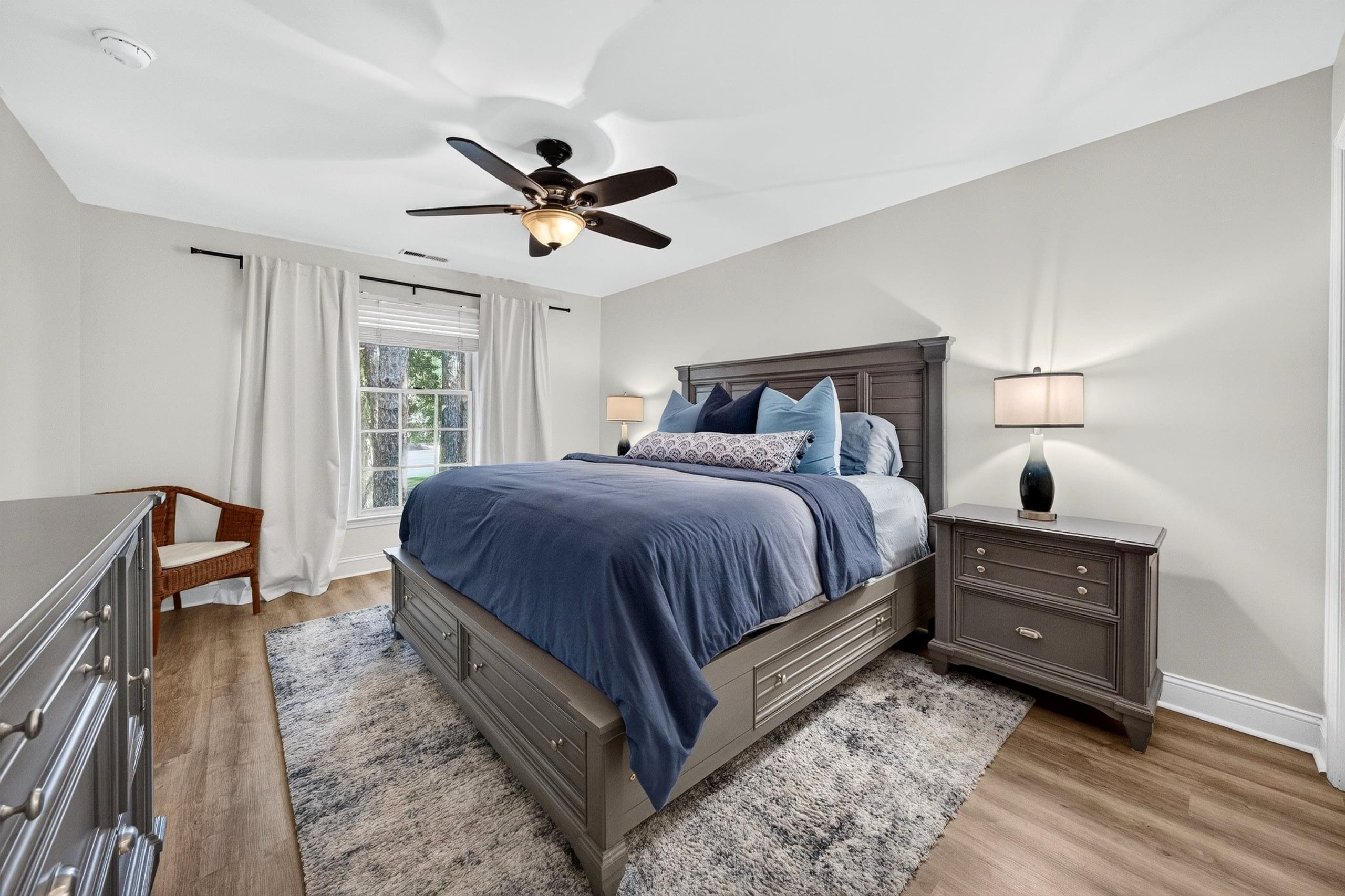 A bedroom with a bed, nightstands, a dresser, a ceiling fan, and a window with curtains.