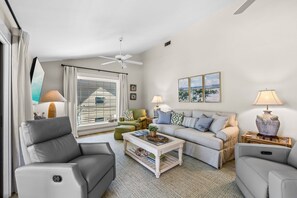 Ein Wohnzimmer mit grauem Sofa, einem grünen Sessel, einem Couchtisch und einem Deckenventilator.
