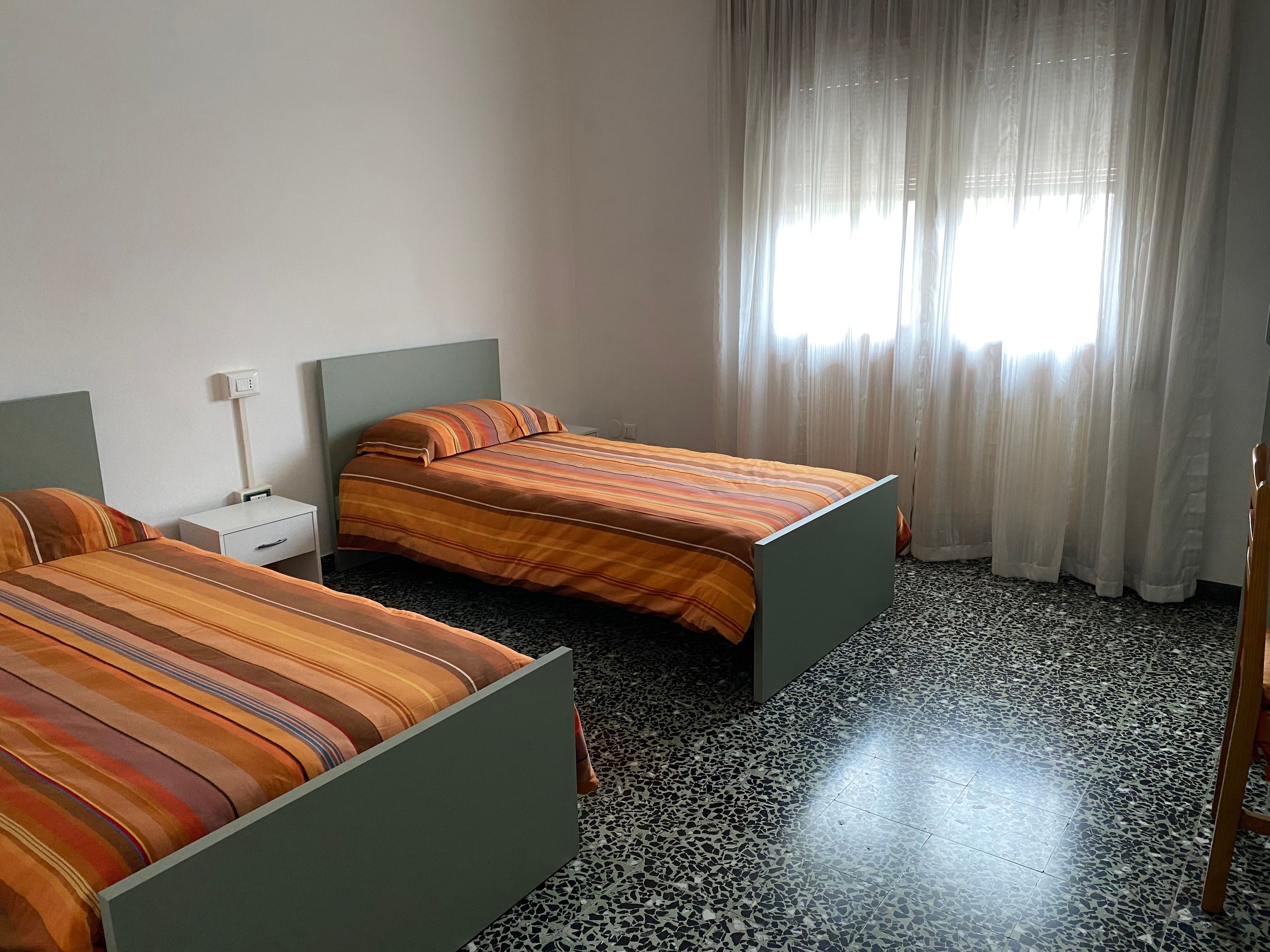 Ein Hotelzimmer mit zwei Betten, einem Nachttisch, einer Steckdose und einem Fenster mit durchsichtigen Vorhängen.