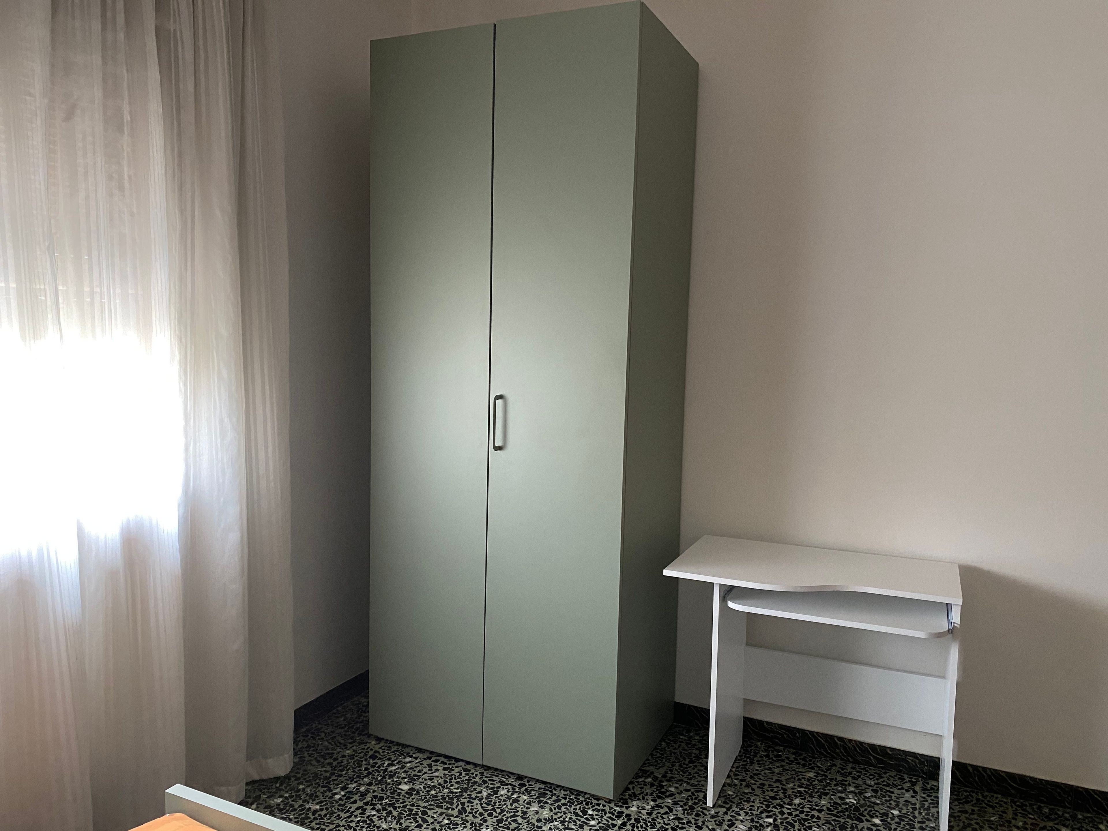 Ein Zimmer mit Kleiderschrank, Schreibtisch und einem Vorhangfenster.