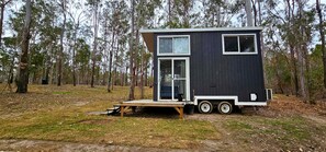 A small, dark-colored cabin with a sliding door and a small porch, surrounded by a forest.