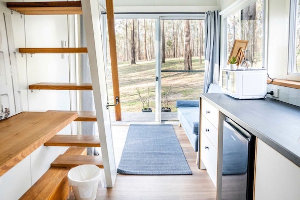 A modern kitchen with wooden shelves, a dining area, and a view of a forest through a glass door.
