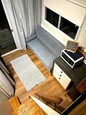 A compact room with a grey sofa, a small white rug, a wooden floor, a microwave on a wooden cabinet, and a sign on the microwave.