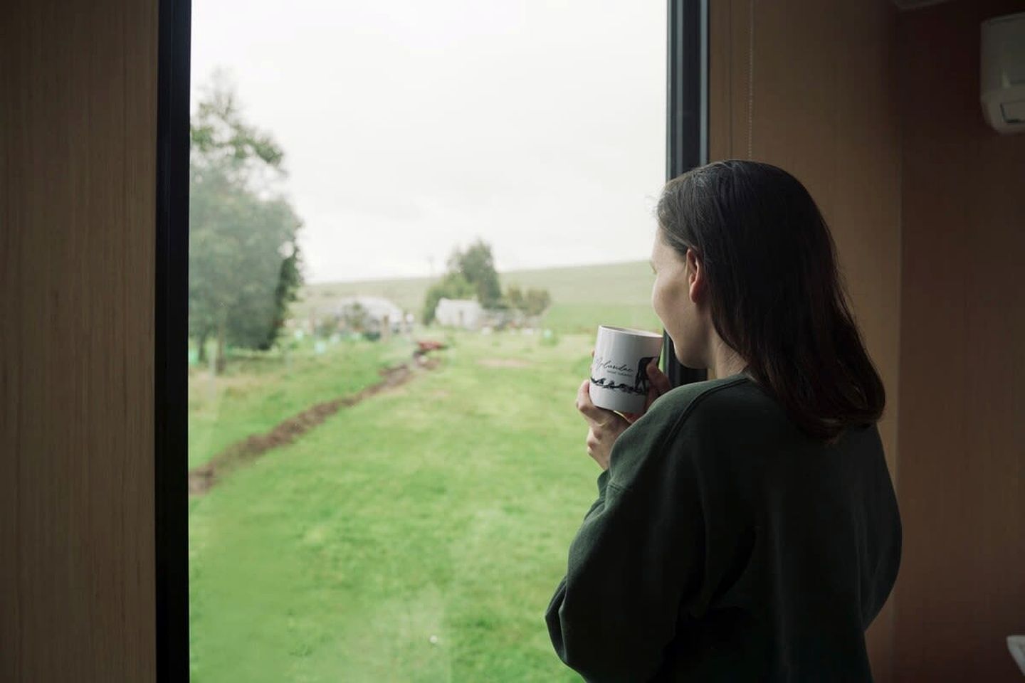 Une personne tenant une tasse, debout près d’une fenêtre donnant sur un champ verdoyant et des arbres.