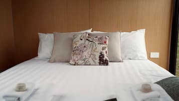 A neatly made bed with white linens and a floral pillow, a wooden headboard, and a window with a view.