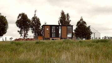 A modern, single-story cabin with a large glass door and windows, surrounded by tall trees and a grassy field.