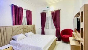 A hotel room with a bed, a nightstand, a red chair, a TV, and a window with curtains. - Paris Arena Holiday Inn (Abuja)