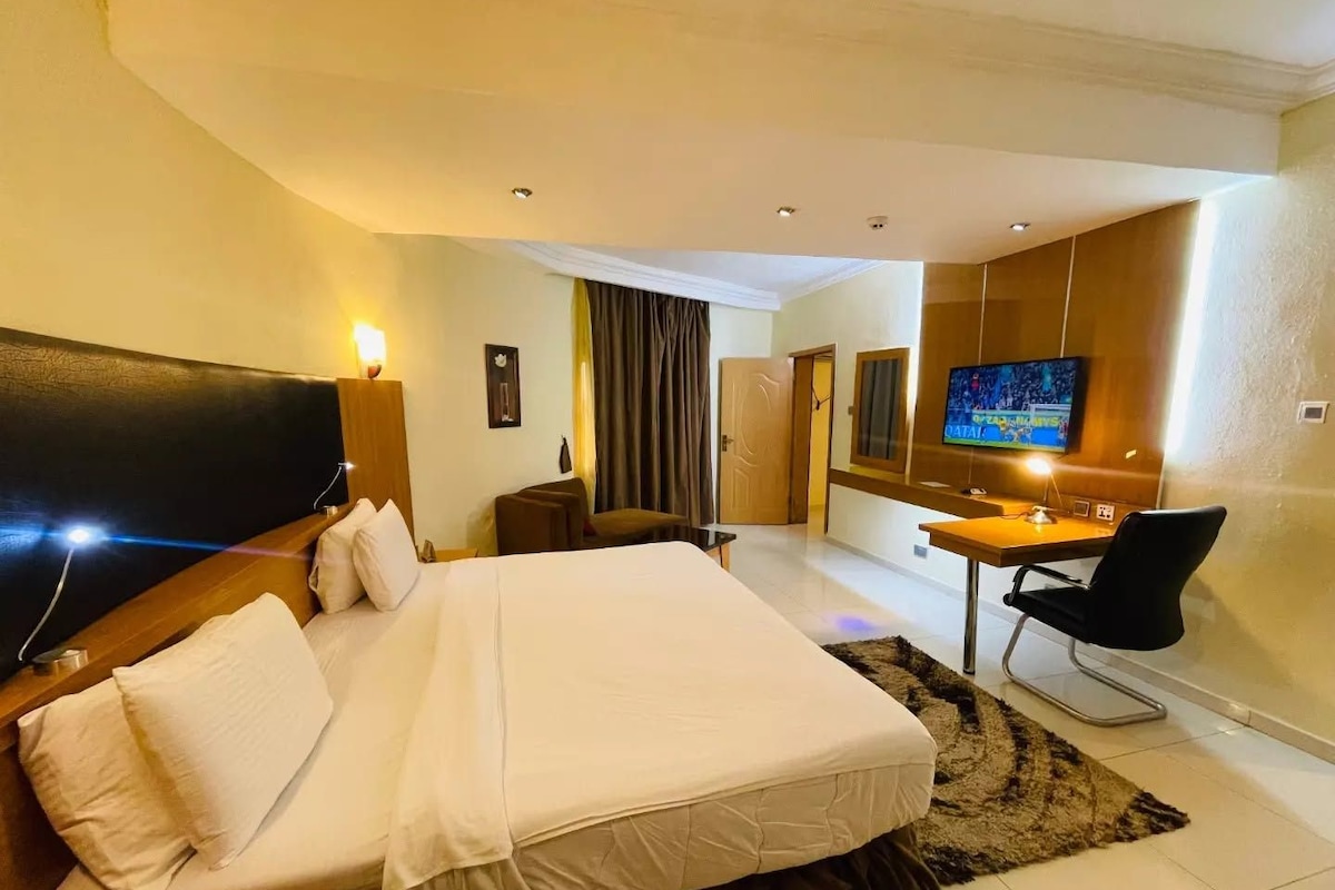 A hotel room with a large bed, a desk with a chair, a television, and a door leading to another room.