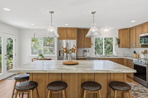 Une cuisine moderne dotée d’un grand îlot central, de meubles en bois, d’appareils électroménagers en acier inoxydable, de luminaires suspendus et offrant une vue sur la verdure environnante.