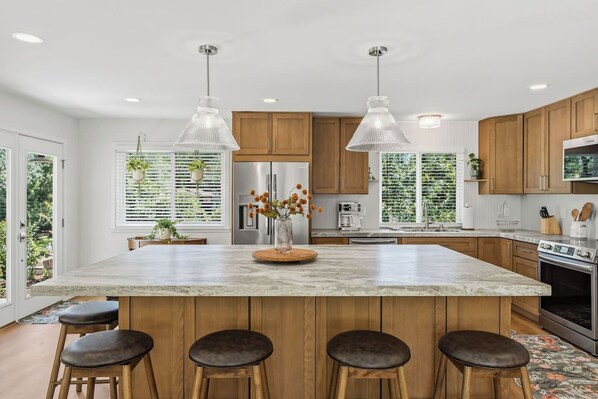 Une cuisine moderne dotée d’un grand îlot central, de meubles en bois, d’appareils électroménagers en acier inoxydable, de luminaires suspendus et offrant une vue sur la verdure environnante.