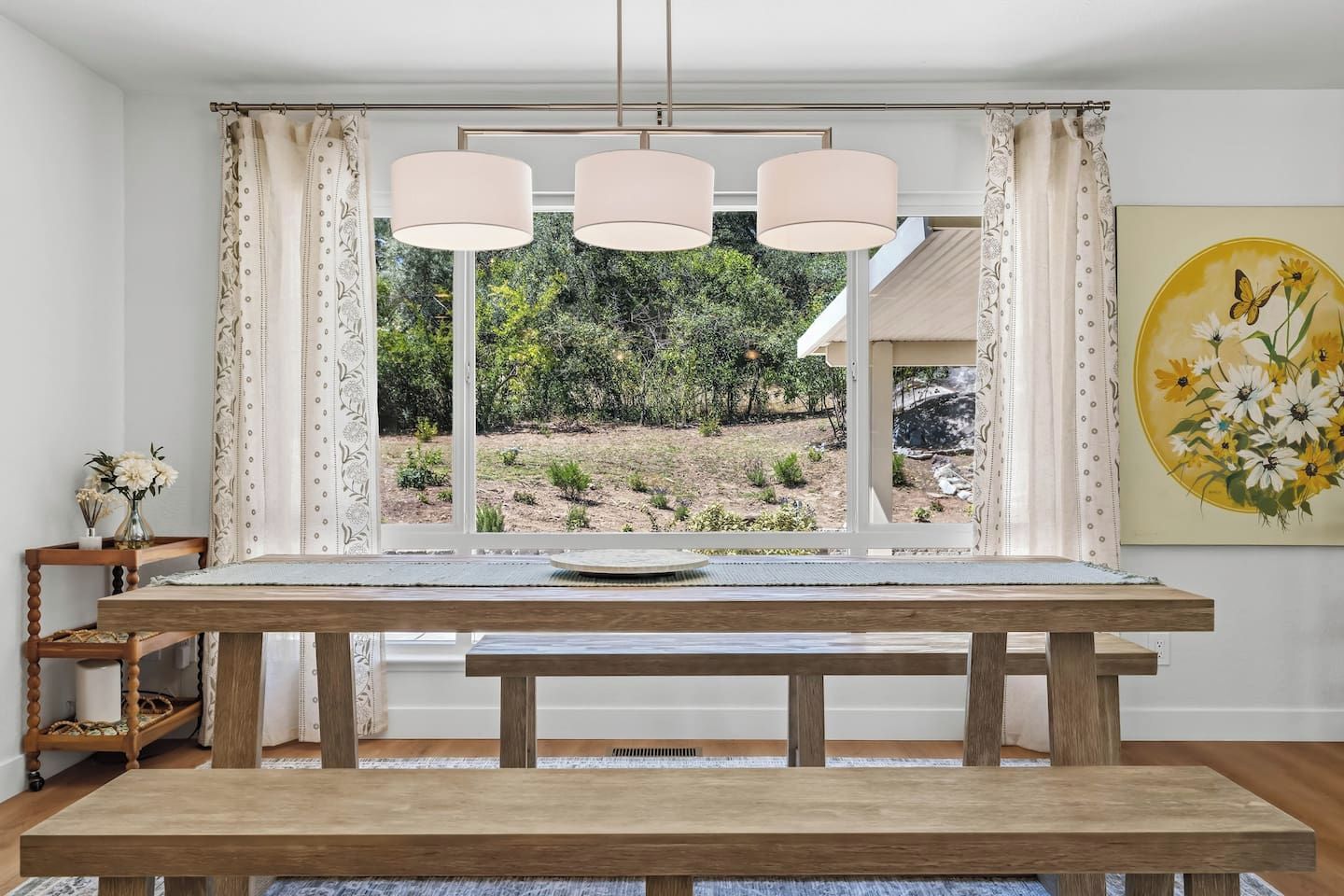 A dining area with a wooden table, three pendant lights, and a view of a garden through a window.