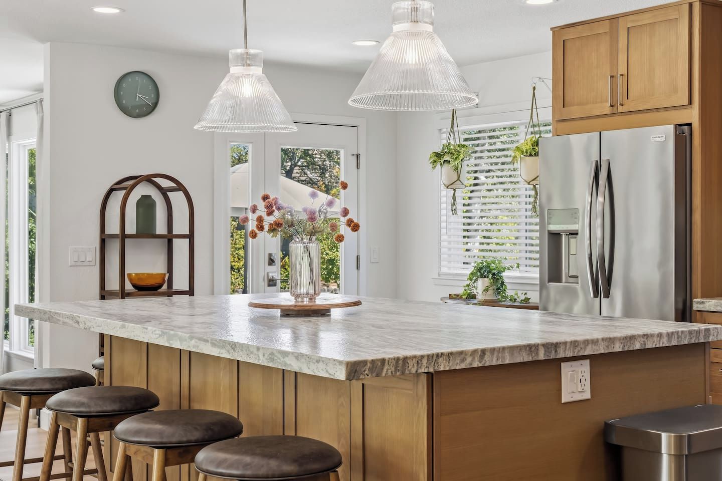 A modern kitchen with a marble countertop, wooden stools, pendant lights, and a built-in refrigerator.