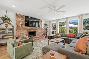 A modern living room with a brick fireplace, a flat-screen TV, and a large window offering a view of the outdoors. - Stunning 4BR Estate | Secluded Folsom Getaway (Folsom)