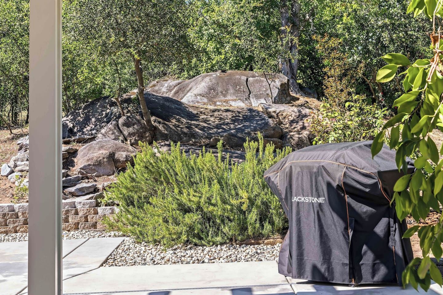 Un espace patio doté d’un barbecue recouvert d’une housse de protection à l’effigie de la marque, entouré de rochers et de verdure.