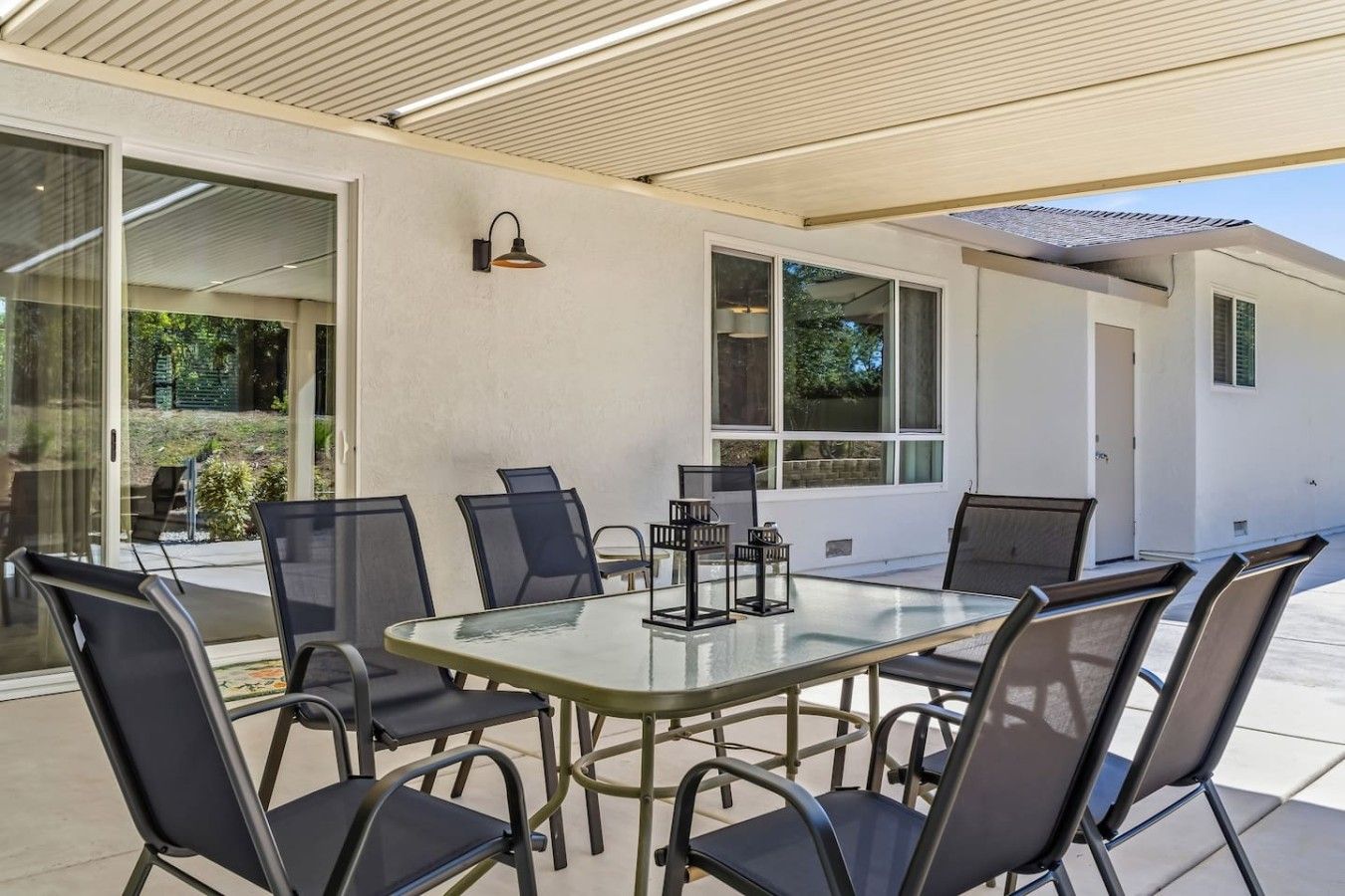 An outdoor patio with a glass table surrounded by chairs, a lamp, and a view of a garden.