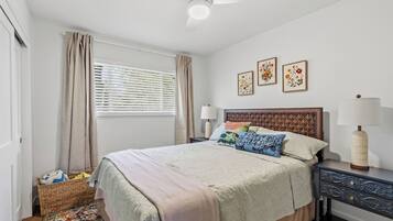 A bedroom with a bed, a nightstand, a lamp, a window with curtains, and a wall with framed pictures.