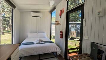 A bedroom with a bed, a small table, a fire extinguisher, and a view of the outdoors.