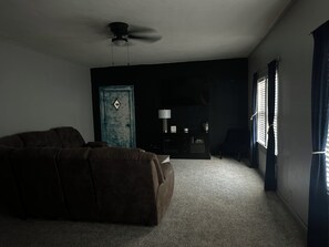 Una habitación con poca luz, un sofá marrón, un ventilador de techo, una puerta y una ventana con cortinas.