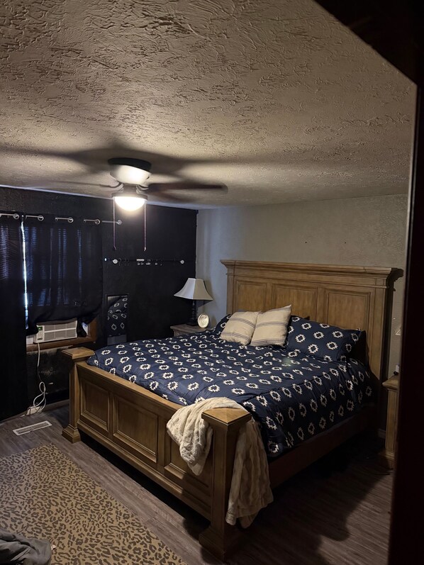Un dormitorio con una cama de madera, un ventilador de techo, una mesita de noche con lámpara y una ventana con cortinas.
