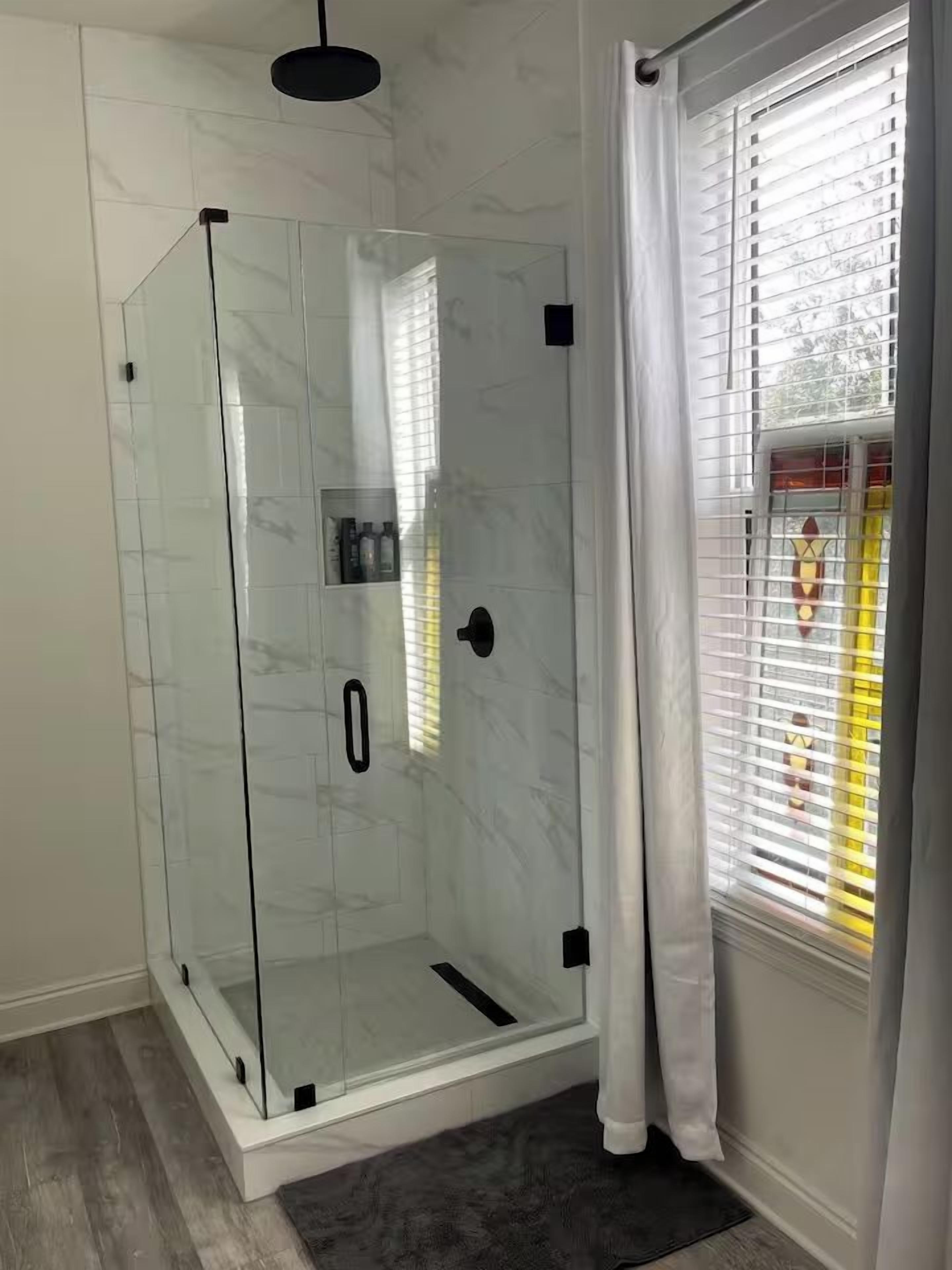 A glass-enclosed shower with a door, a window with blinds, and a dark rug on the floor.